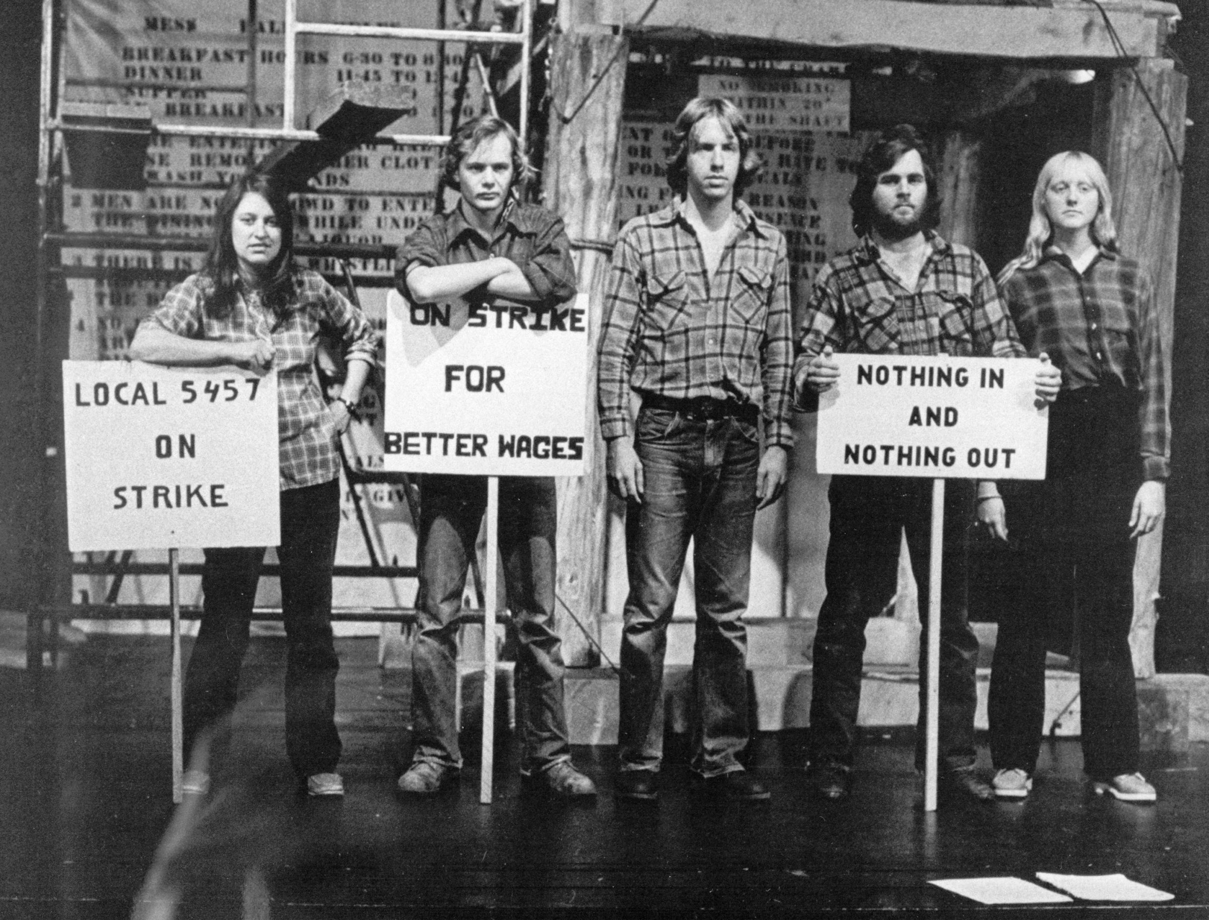A black and white photo of 5 people with 3 strike signs in front of them. They're each wearing jeans and plaid shirts.