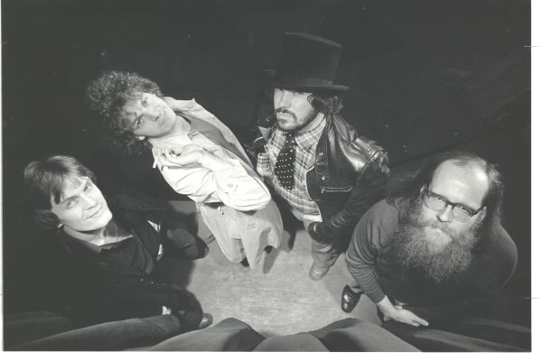 A black and white photo of four people looking up at the camera. The first person is wearing a black coat, the second a white jacket and is holding a cigarette. The third is wearing a top hat, plaid shirt under a leather jacket and a tie. The fourth has a large beard and glasses and long sleeved shirt.
