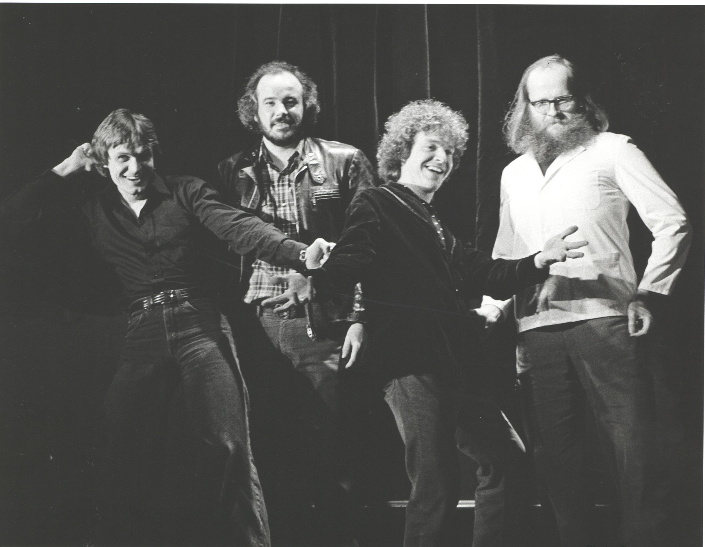 A black and white photo of four people. The first is wearing a black collared shirt and jeans, the second a plaif shirt under a leather jacket and jeans, the third a black coat and jeans, and the fourth has a large beard and glasses and is wearing a white shirt and jeans. The first person is pusing on the third persons arm. Everyone is smiling.
