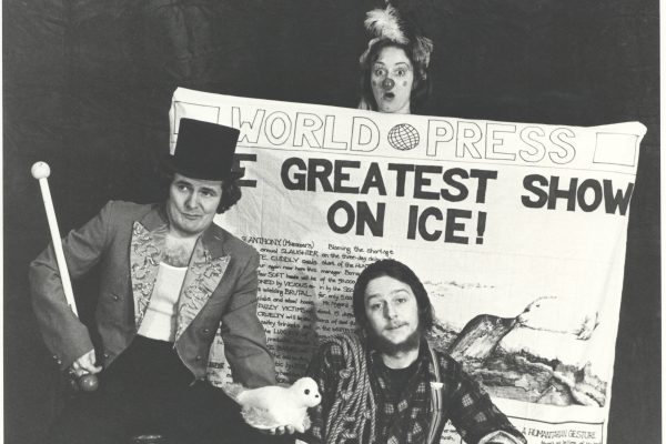 A black and white photo of three people. The first is wearing a top hat, circus blazer and is holding a stuffed seal toy. A person with a beard and wearing a plaid shirt has rope over their shoulder is sitting next to them. A person ith feathers in their hair and dressed as a clown is holding a large sheet with World Press Greatest Show on Ice! behind them.