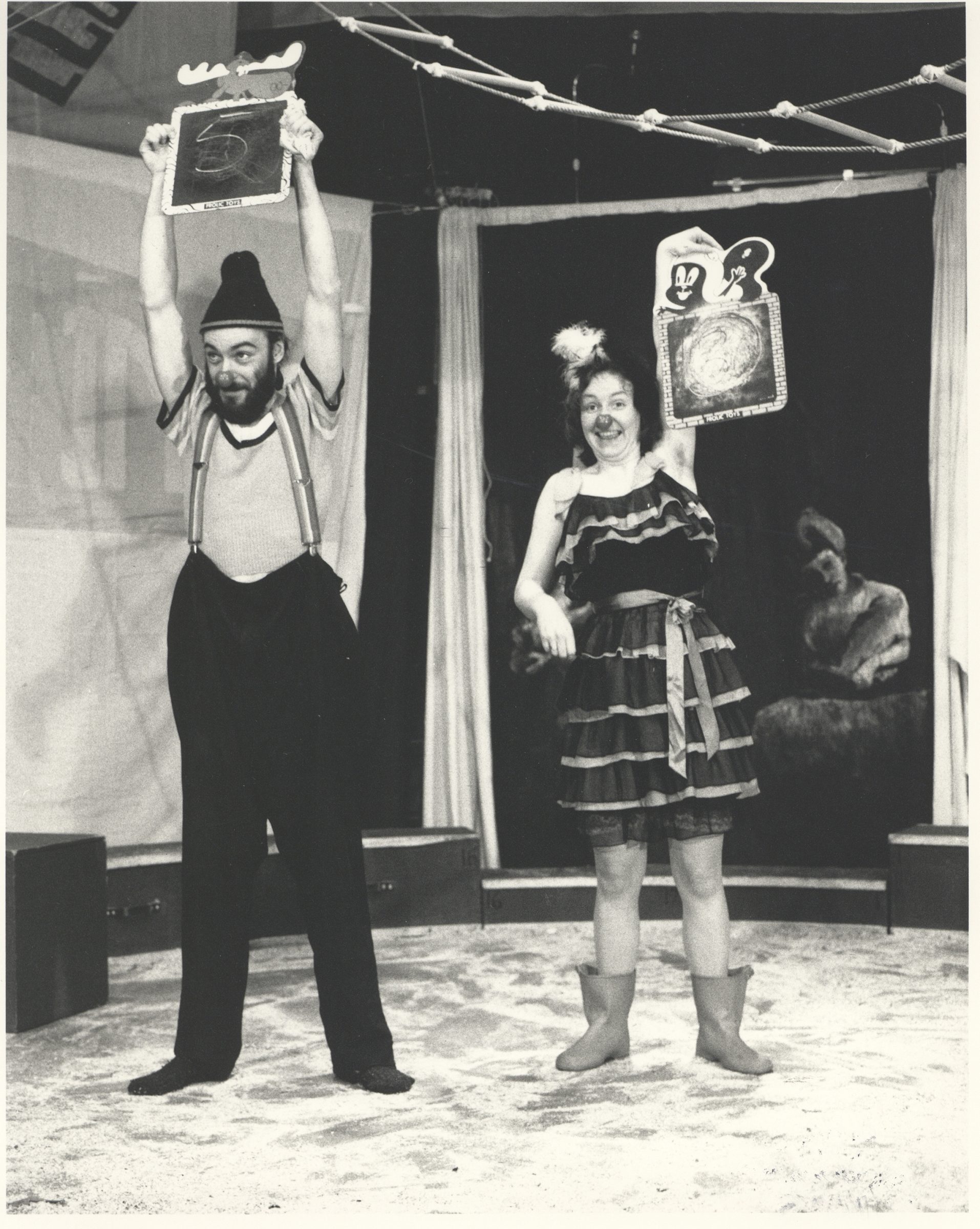 A black and white photo of two people on stage. The first is wearing a touque and suspenders holding up black pants. The person is holding a chaulkboard above their head with a 5 on it. The second person is wearing a dress, rubber boots and a clown nose. Their holding a chaulkboard with a circle.