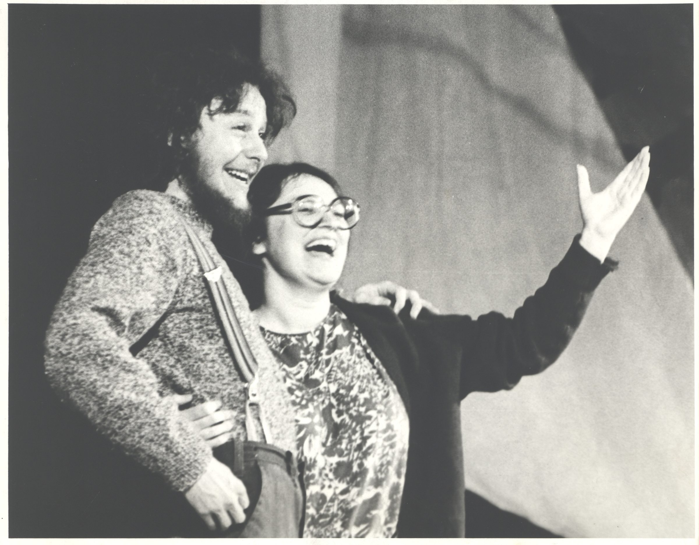 A black and white photo of two people holding each other side by side. They are both smiling and one is gesturing to the audience with their arm up.