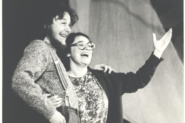 A black and white photo of two people holding each other side by side. They are both smiling and one is gesturing to the audience with their arm up.