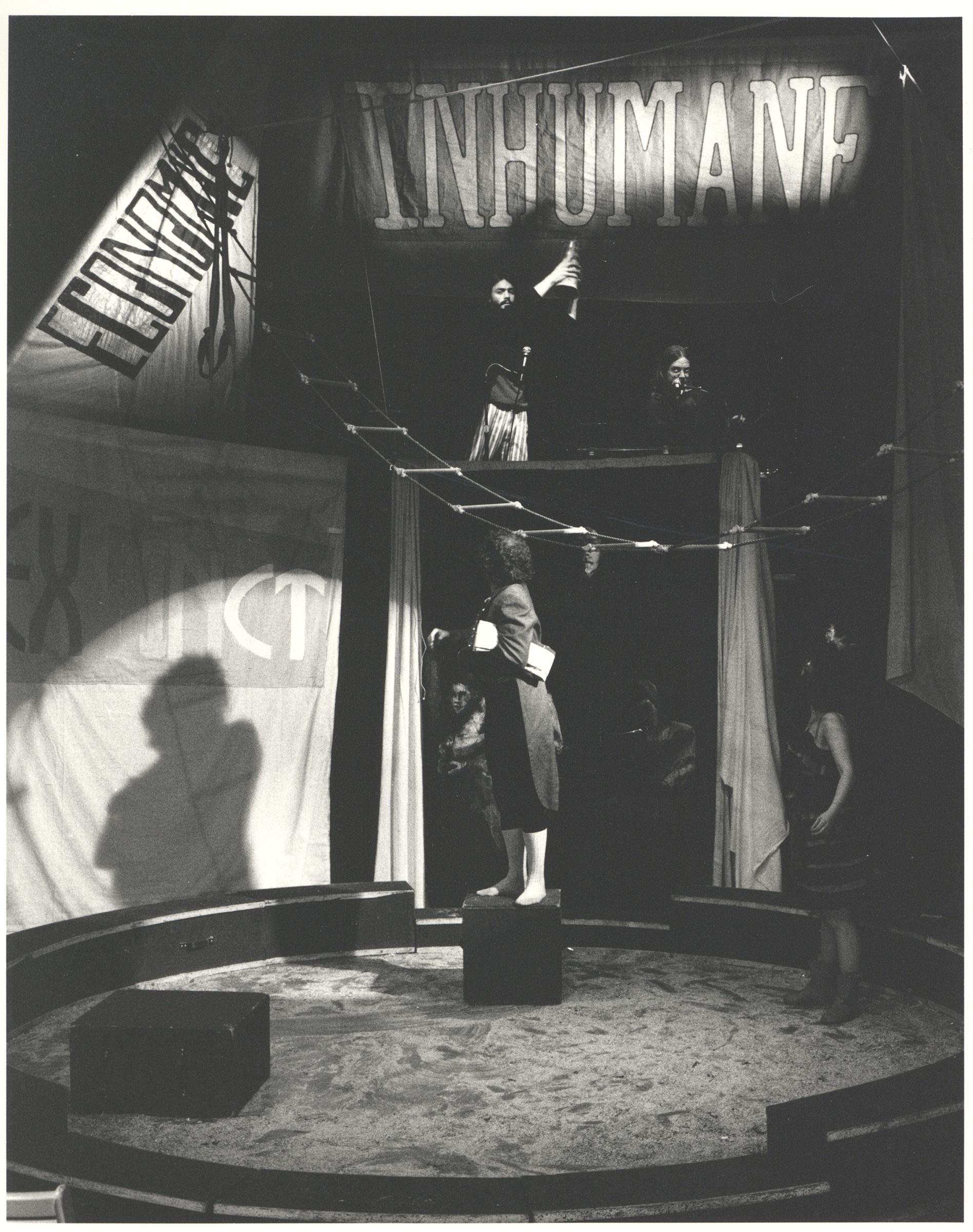 A black and white photo of a circus ring. A person stands on a box looking up above them. They're wearing a long jacket. A person on stands to the right wearing a dress, also looking up. Two people are on a platform above them.