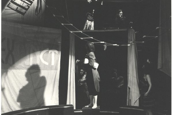 A black and white photo of a circus ring. A person stands on a box looking up above them. They're wearing a long jacket. A person on stands to the right wearing a dress, also looking up. Two people are on a platform above them.
