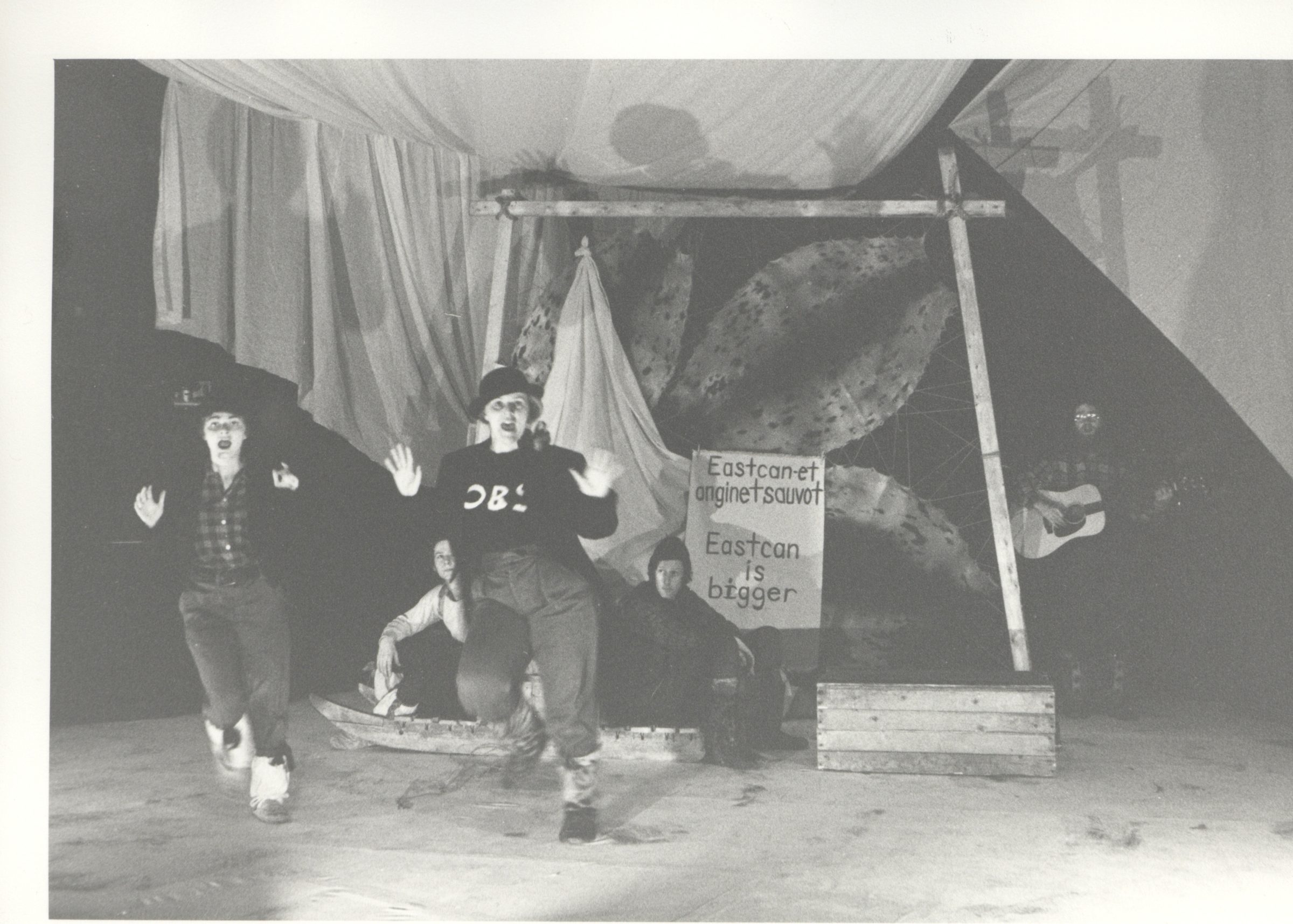 A black and white photo of two people moving with their hands up and mouths open as if singing. Two people sit behind them on a sled. Seal skins are stretched out in a frame behind the sled. A sign says Eastcan-et anginetsauvot Eatcan is bigger.