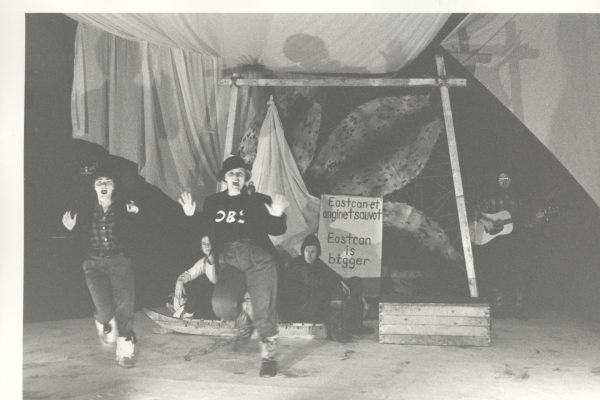 A black and white photo of two people moving with their hands up and mouths open as if singing. Two people sit behind them on a sled. Seal skins are stretched out in a frame behind the sled. A sign says Eastcan-et anginetsauvot Eatcan is bigger.