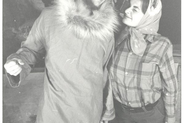 A black and white photo of two people. One is wearing a fur collared coat and boots and smiling down at t person wearing a scarf around their head, a plaid shirt and boots. They are taking a picture of the taller person.