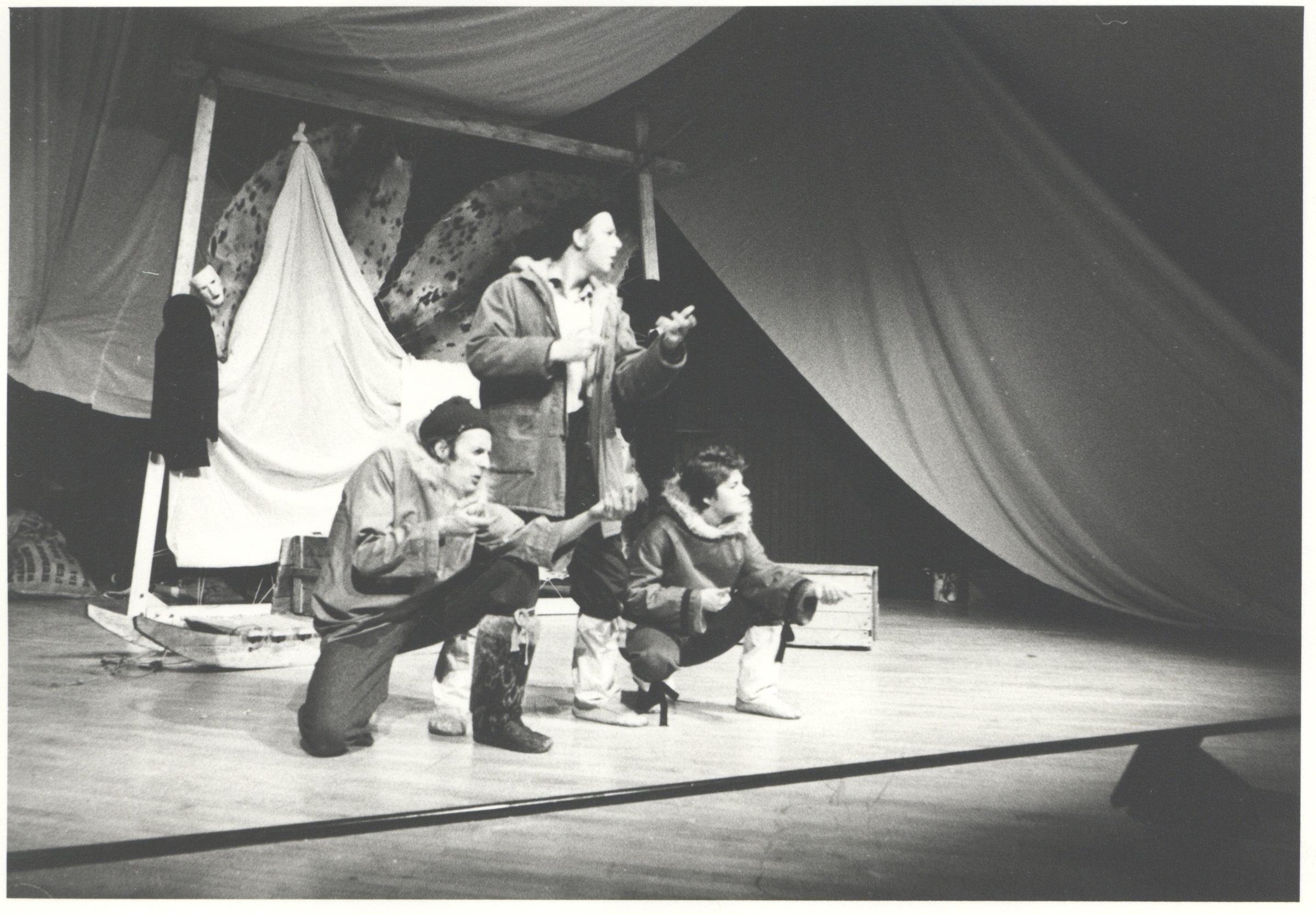 A black and white photo of three people on stage all gesturing to the audience. Two are crouched down and one is standing. They're all wearing winter clothes. Seal skins are stretched out behind them.