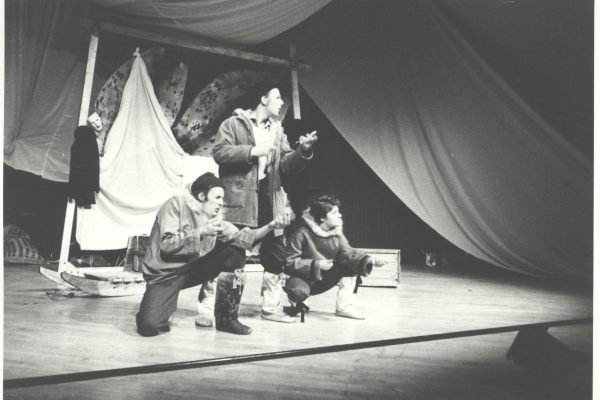 A black and white photo of three people on stage all gesturing to the audience. Two are crouched down and one is standing. They're all wearing winter clothes. Seal skins are stretched out behind them.