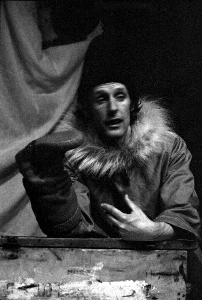 A black and white photo of a person wearing a fur collared coat and dark touque. They are gesturing and holding a large slipper boot on their hand.