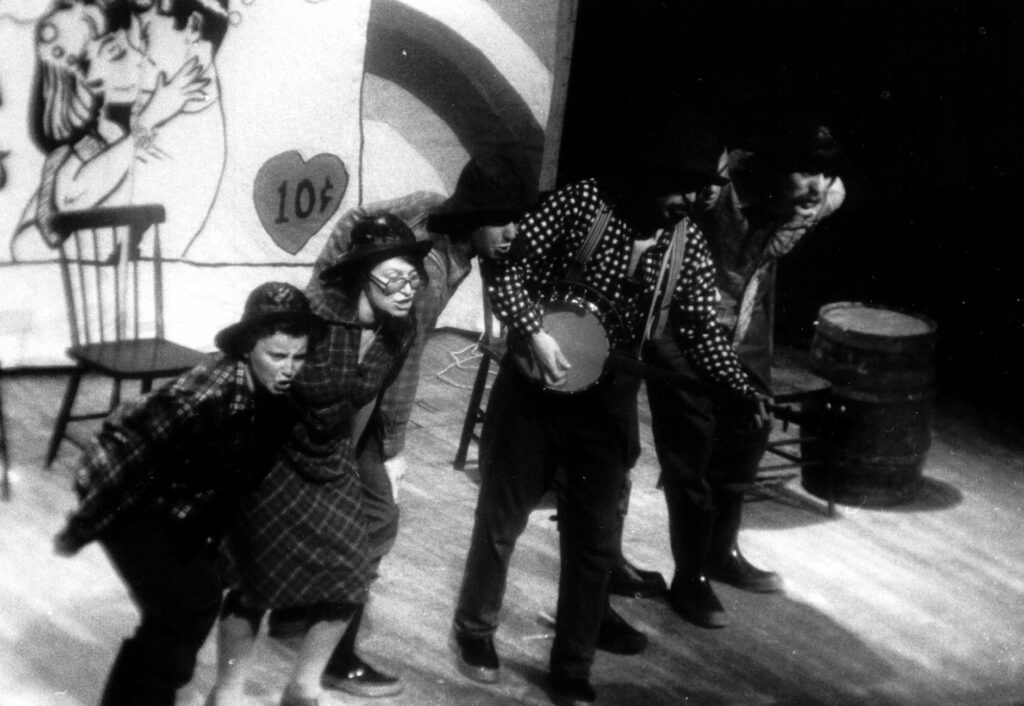 Five people in a row on stage. They are all bent at the waist leaning towards the audience singing. The fourth person in line is playing a banjo. Wooden chairs and a barrel are behind them. The back wall has a comic of people kissing and a large 10 cents in a heart.