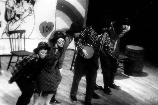 Five people in a row on stage. They are all bent at the waist leaning towards the audience singing. The fourth person in line is playing a banjo. Wooden chairs and a barrel are behind them. The back wall has a comic of people kissing and a large 10 cents in a heart.