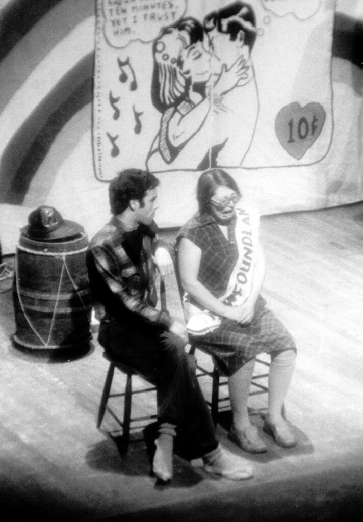 A black and white photo of two people on stage sitting on wooden chairs. One person is crying and wearing a dress and a large white sash with Newoundland written across it. The other person is wearing jeans and a plaid shirt and facing the crying person.