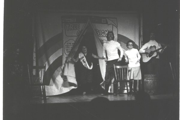 A black and white photo of four people on stage. One is coming through curtains in the middle of the stage and crying, wearing a sash with Newfoundland on it. The three other people are wearing white sweaters and singing. One person is playing guitar.