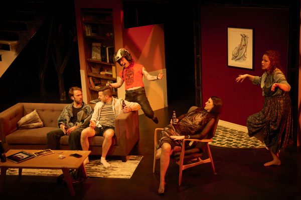 A group of young adults hang around a living room set. Two men on the left are sitting on a couch. One woman stands on the right, with another woman sitting on a chair in the middle. Behind her, a man in the background wears a red shirt and motorcycle helmet with his arms held out wide.