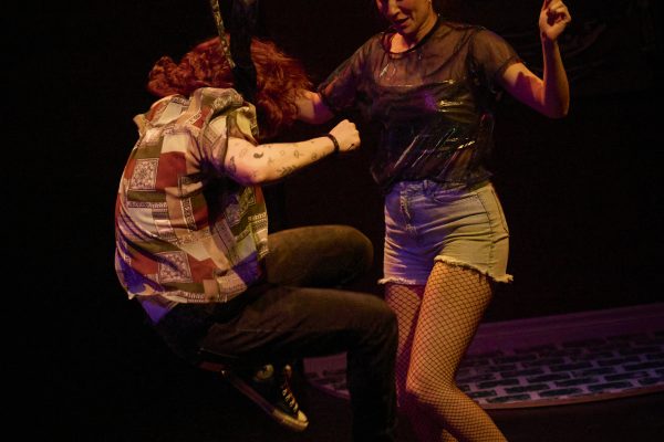 A young women wearing a black t-shirt, jean shorts, and fishnet tights dances with a man wearing a patterned shirt and dark pants.