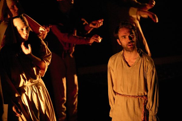 A young man in beige clothing stands and looks out at the audience. A group of people in beige clothing dance around him.