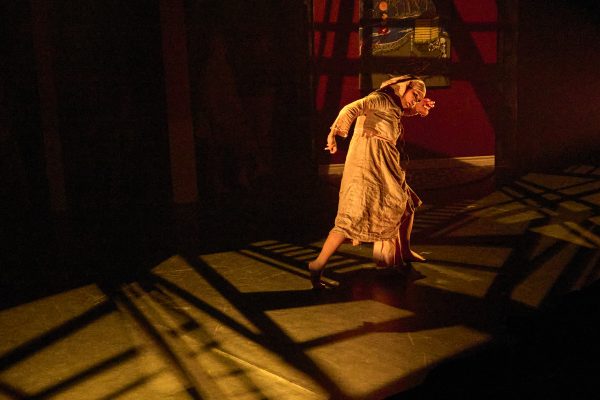 A young woman in beige clothing dances in front of a black background. Long shadows are cast across her and the stage floor.