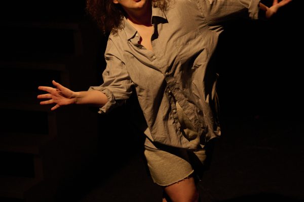 A young woman with red hair and beige clothing dances in front of a black background.